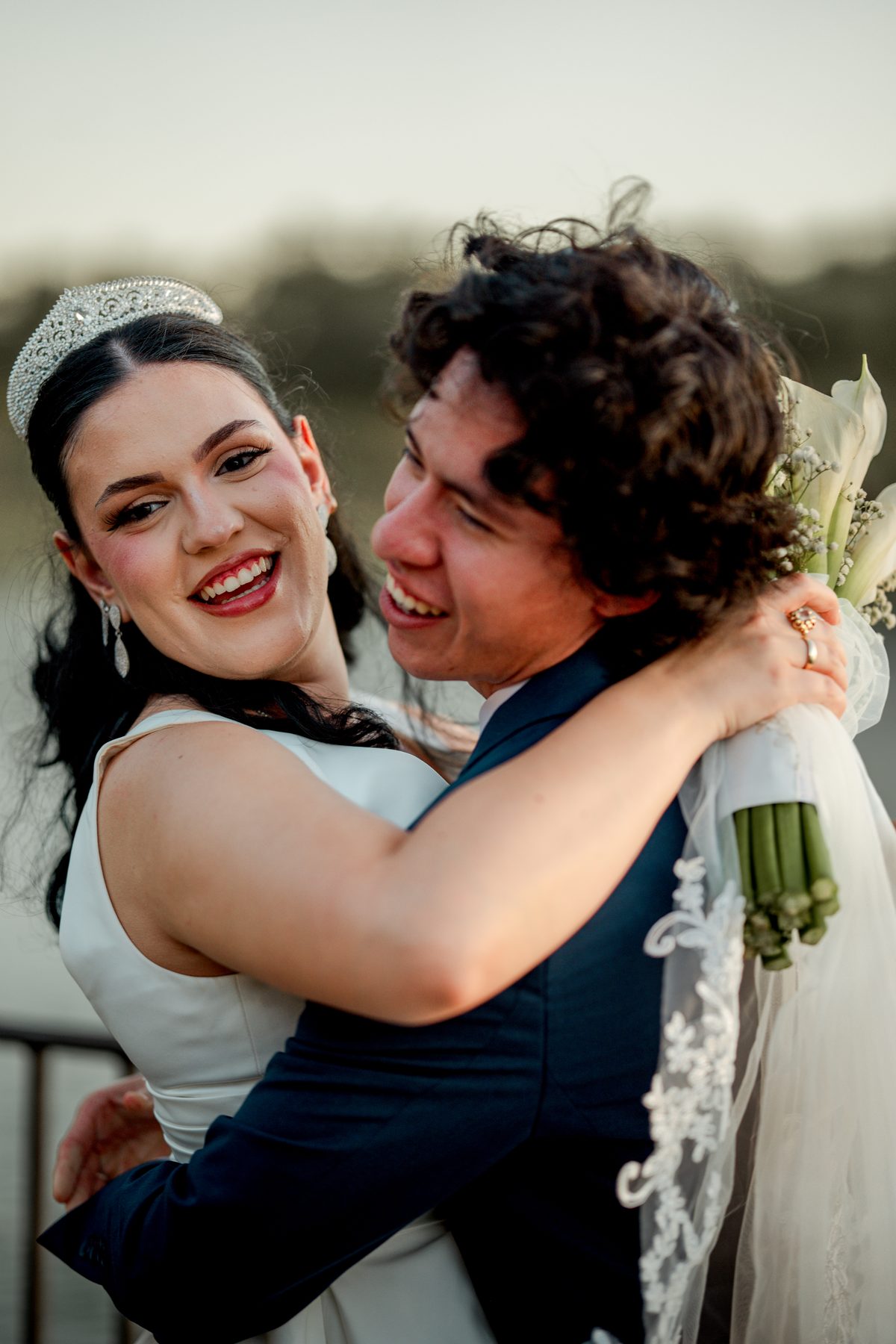 A bride in a tiara laughing as the groom lifts her, bouquet in hand
