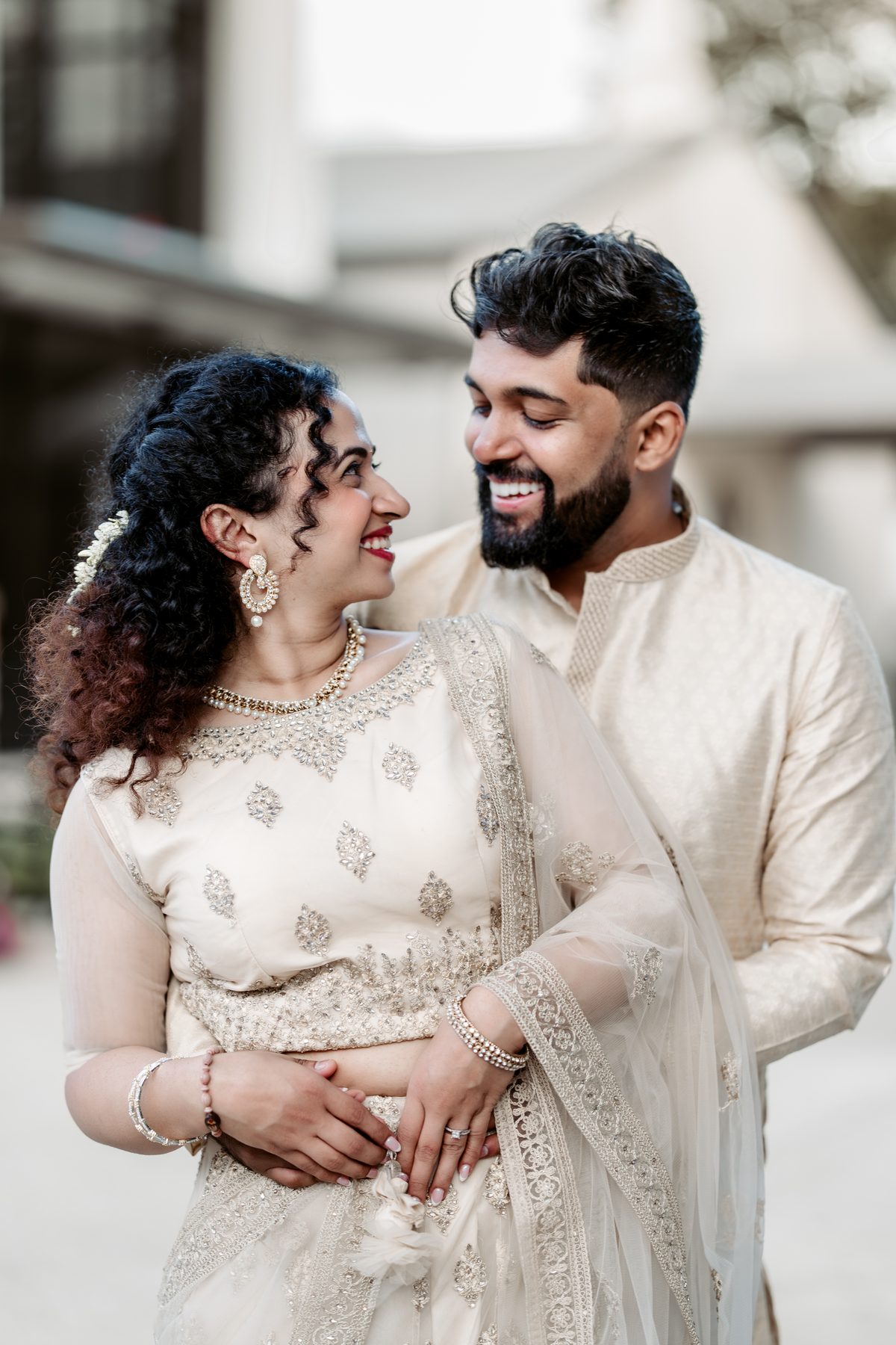 A couple in cream and gold, laughing, in front of a house